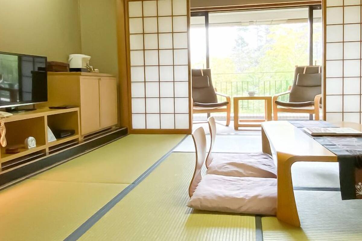 A traditional Japanese-style room with tatami flooring, a television, a cabinet, and a sliding door leading to a balcony with chairs.