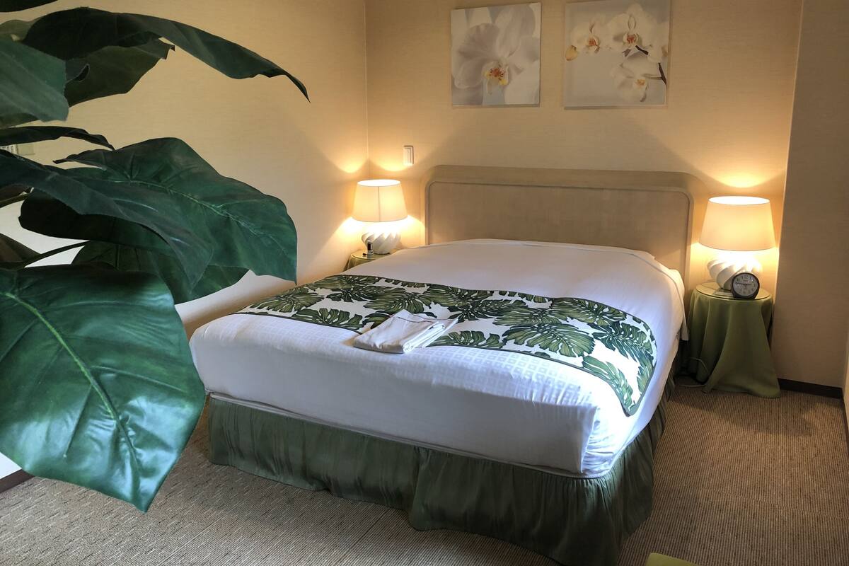 A hotel room with a bed, two wall-mounted lamps, a large green plant, and two framed floral pictures on the wall.