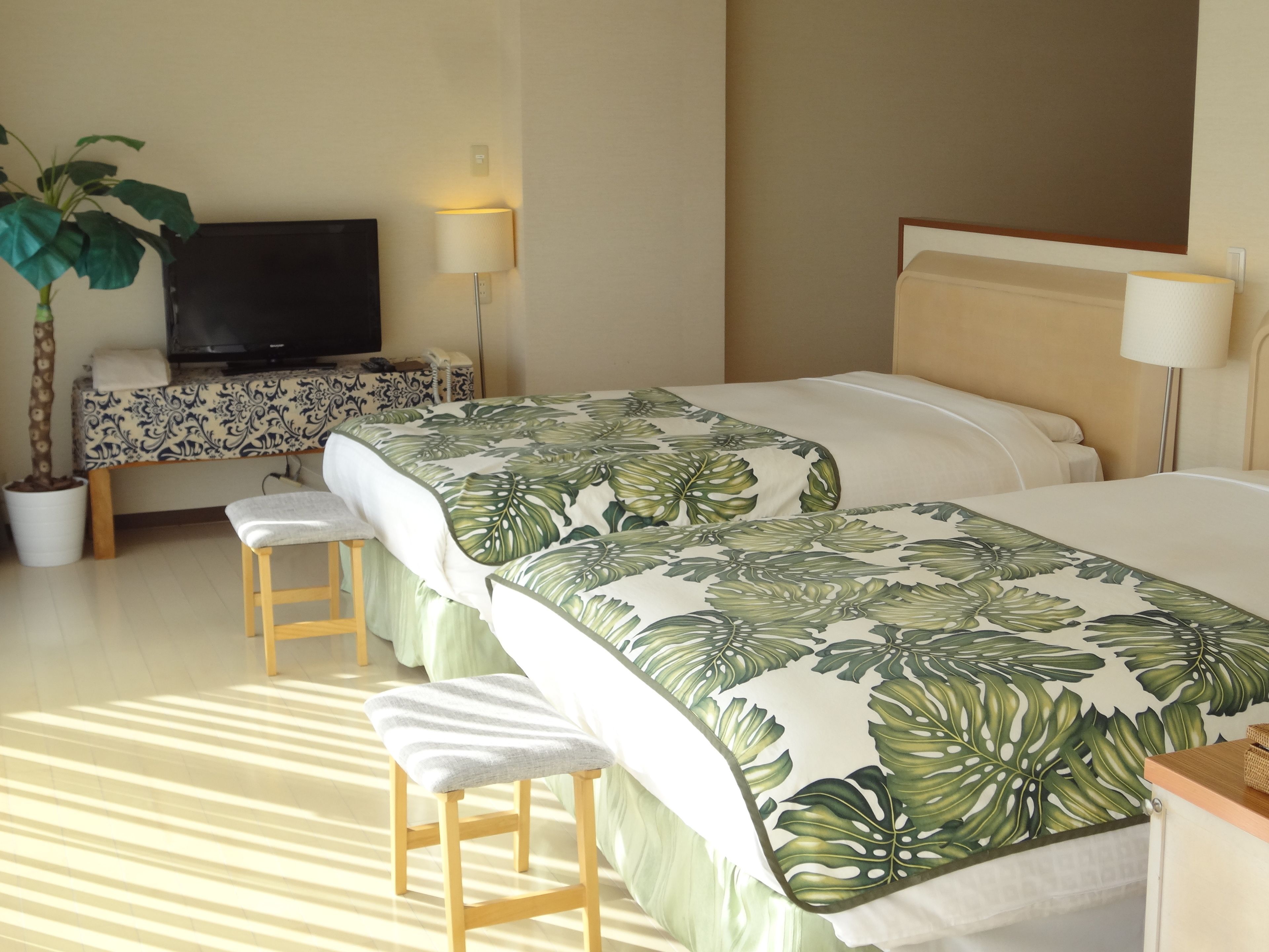 A hotel room with two beds, a TV, a desk, a chair, a potted plant, and a lamp.