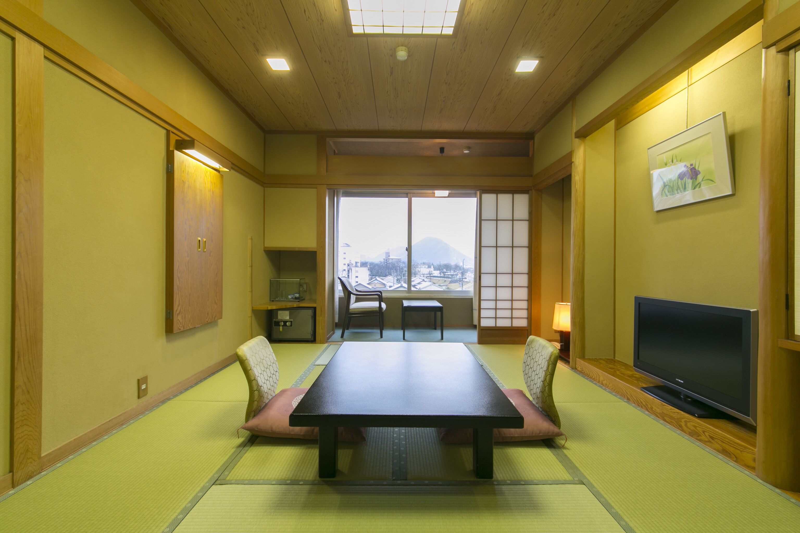 A traditional Japanese room with tatami flooring, a low table, seating, a television, and a view of a mountain.