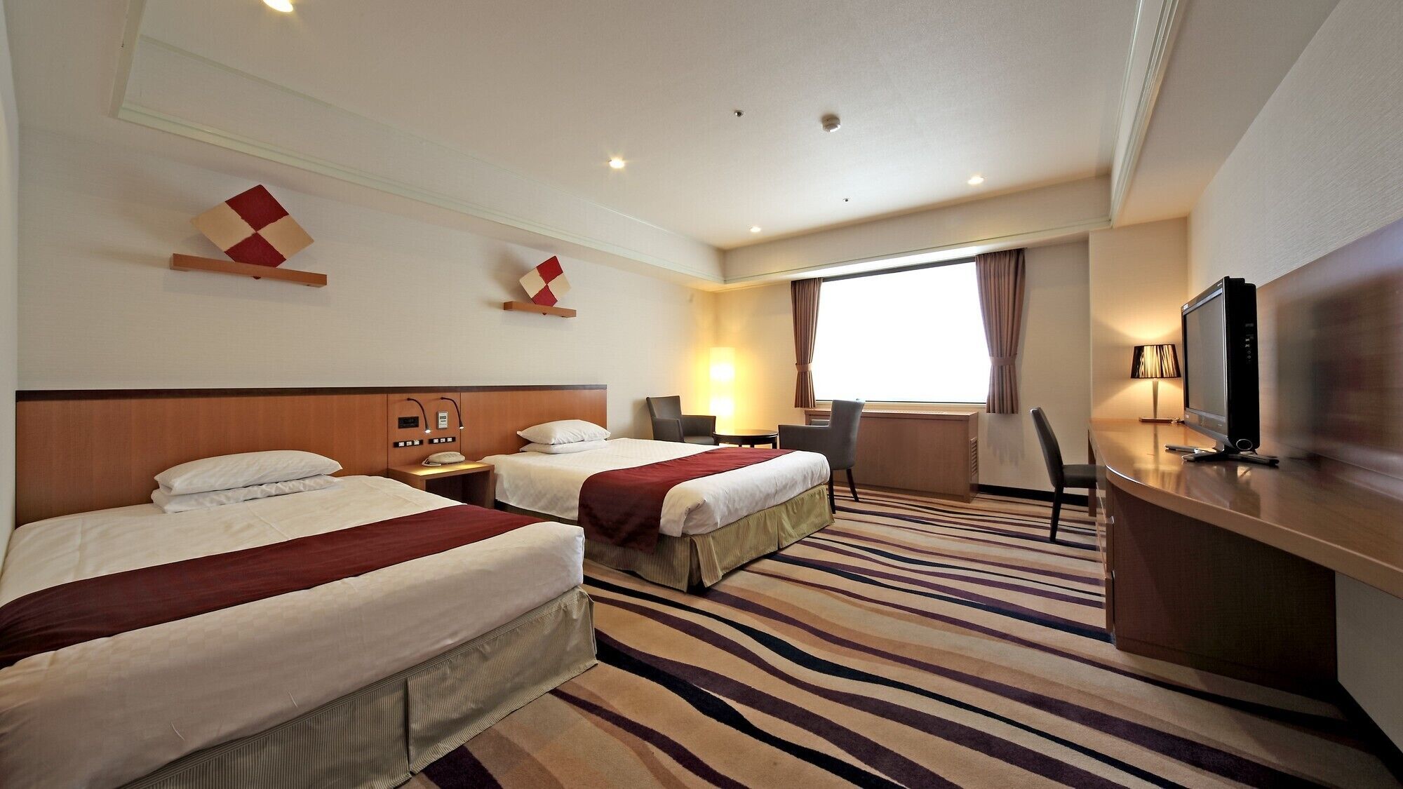 A hotel room with two beds, a desk with a TV, a chair, a lamp, and a window with curtains.