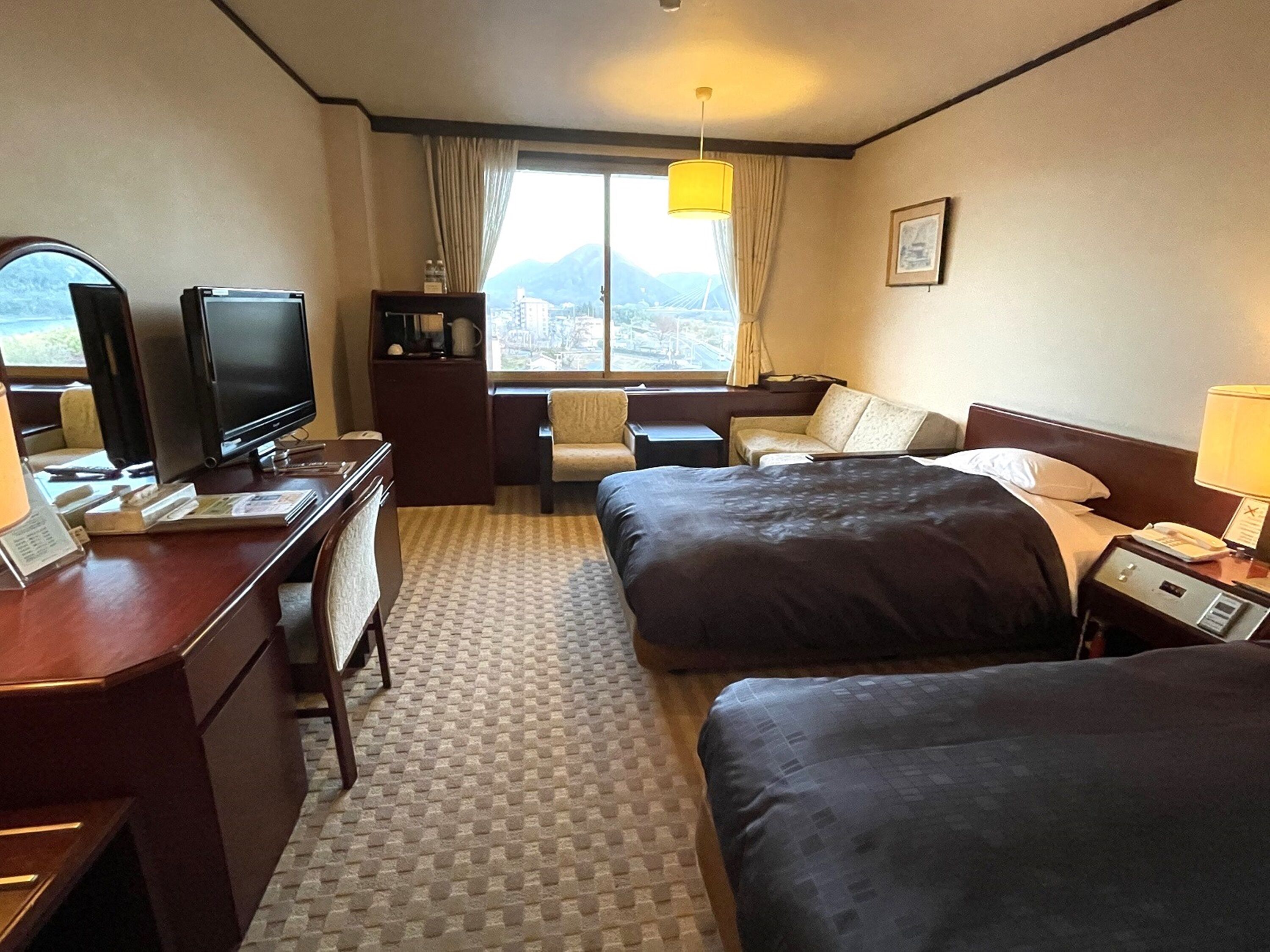 A hotel room with a desk, two beds, a television, a window with a view of mountains, and a lamp.