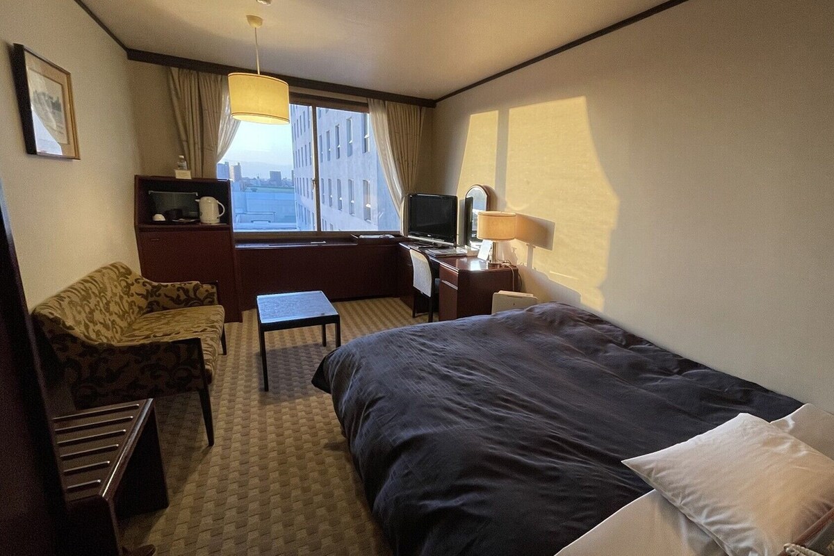 Et hotelværelse med seng, skrivebord, stol, fjernsyn og et vindue med gardiner.