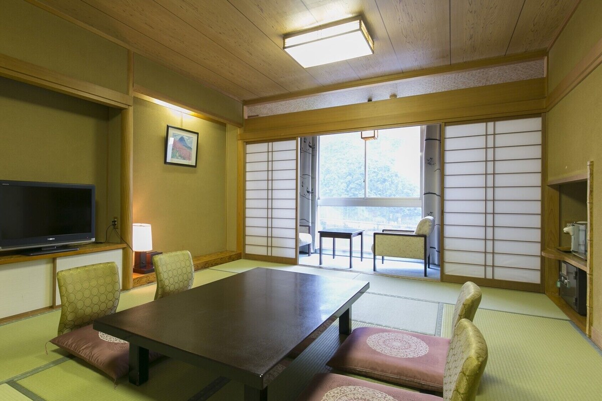 Et traditionelt japansk rum med tatamimåtter, et lavt bord, stole, et fjernsyn og en skydedør til en balkon.