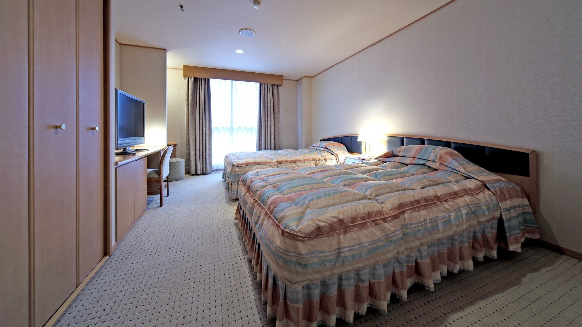 A hotel room with two beds, a desk with a TV, and a large window with curtains.