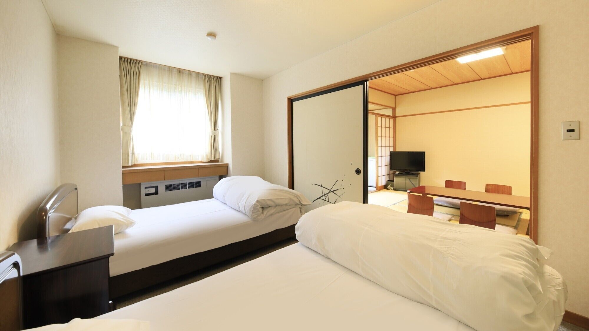 A hotel room with a bed, a desk with a chair, a television, and a sliding door leading to a traditional Japanese room.
