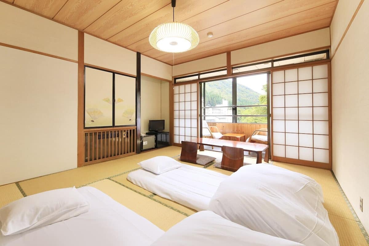 Une pièce de style japonais traditionnel, avec un sol recouvert de tatamis, un plafond bas en bois, des portes coulissantes en papier de riz (shoji), un téléviseur et un balcon offrant une vue sur les montagnes.