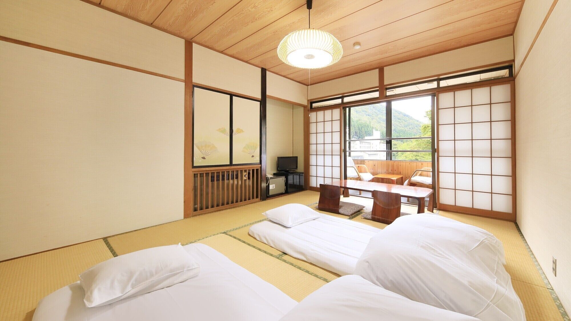 A traditional Japanese room with tatami flooring, a low wooden ceiling, sliding shoji doors, a television, and a view of a mountainous landscape.