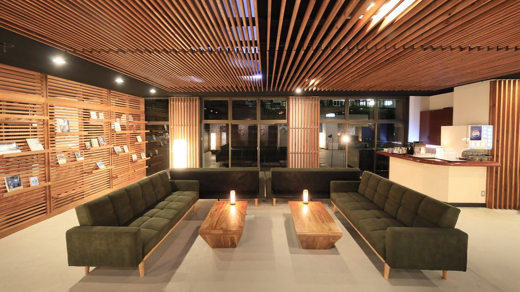 Un hall d’entrée moderne avec un plafond à lambris en bois, des canapés verts et un comptoir de réception équipé d’un distributeur de boissons Pepsi.