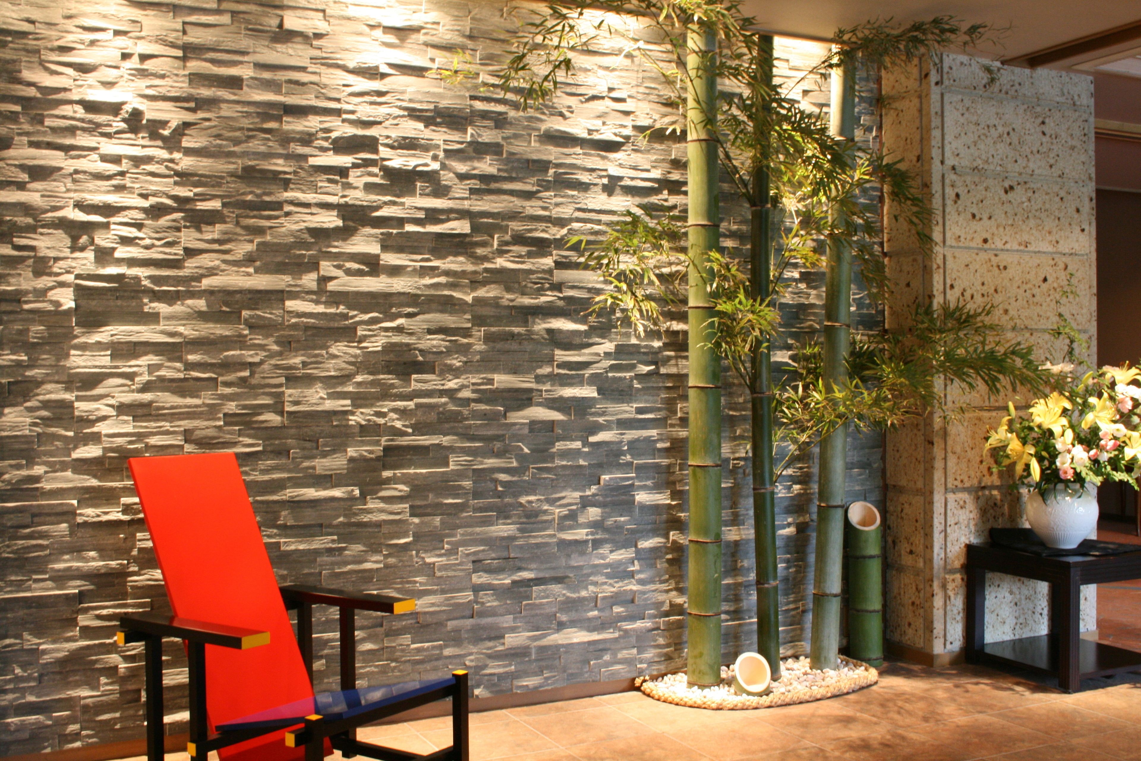 Um interior moderno com uma parede de pedra, uma cadeira preta e vermelha, plantas de bambu e um vaso com flores.