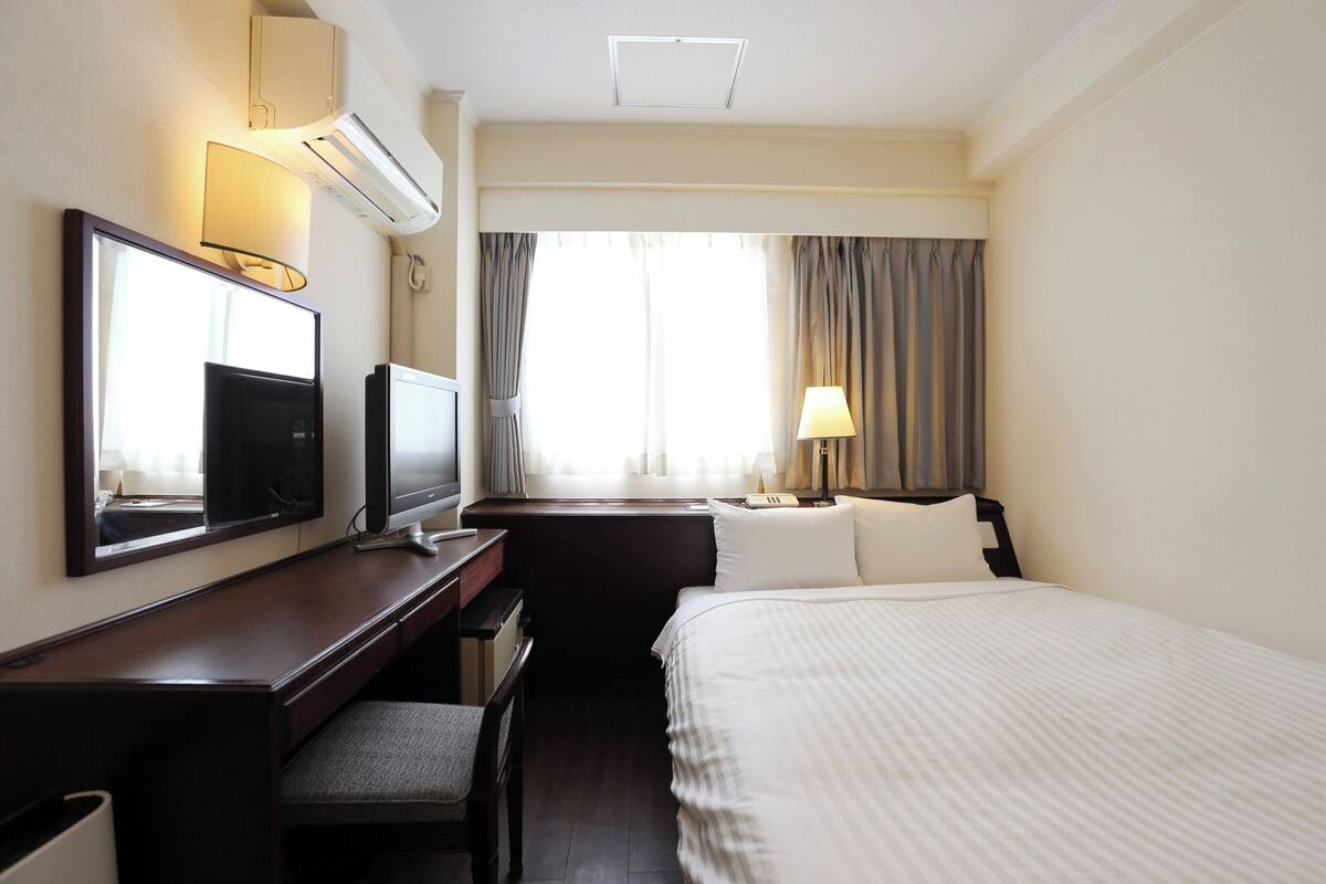 Une chambre d’hôtel avec un lit, un bureau et une chaise, une télévision, une lampe et une fenêtre avec des rideaux.