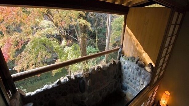 Una bañera de piedra con vista a árboles otoñales, un banco de madera y una lámpara fijada a la pared.