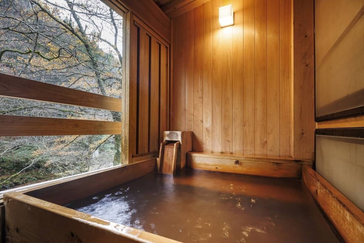 Ein hölzernes Quellbad mit Sitzbank, kleinem Heizgerät und Fenster mit Blick auf Bäume.