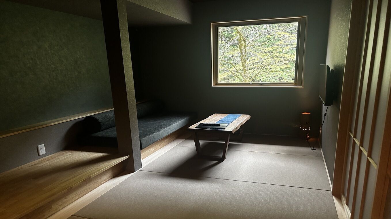 Una stanza con pavimento in tatami, una panca di legno bassa, un tavolino, una finestra con vista sugli alberi e una porta scorrevole tradizionale.