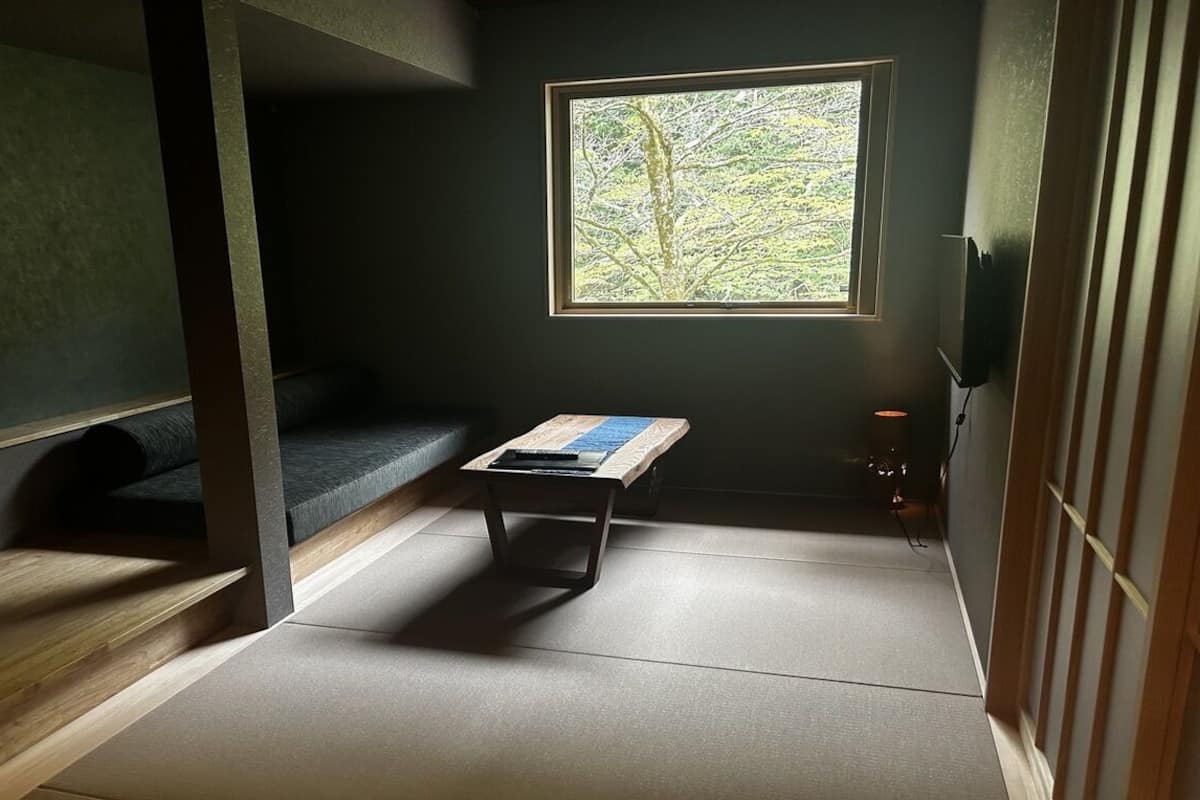 Ein Zimmer mit Tatami-Boden, einer niedrigen Holzbank, einem kleinen Tisch, einem Fenster mit Blick auf Bäume und einer traditionellen Schiebetür.