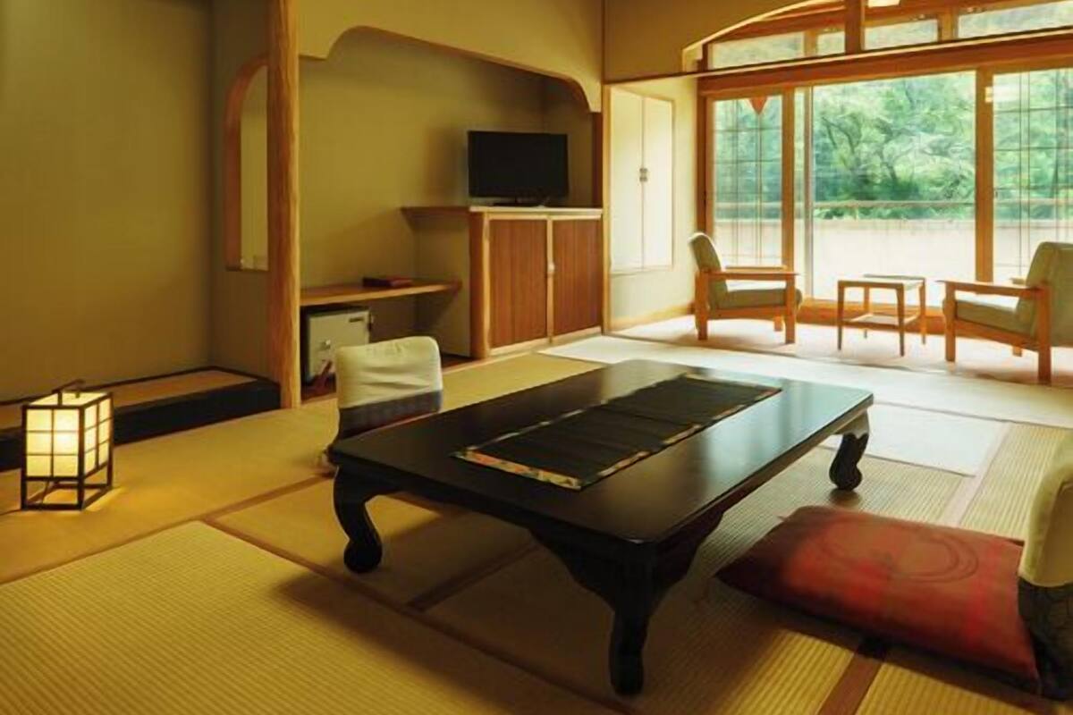 Una habitación tradicional japonesa con suelo de tatami, una mesita baja, televisión y vistas al exterior con vegetación.