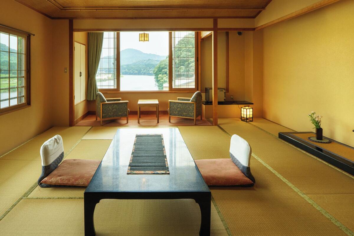 Una habitación japonesa tradicional con suelo de tatami, una mesita baja, dos sillas, vista a un lago y una pequeña mesa con un farol.