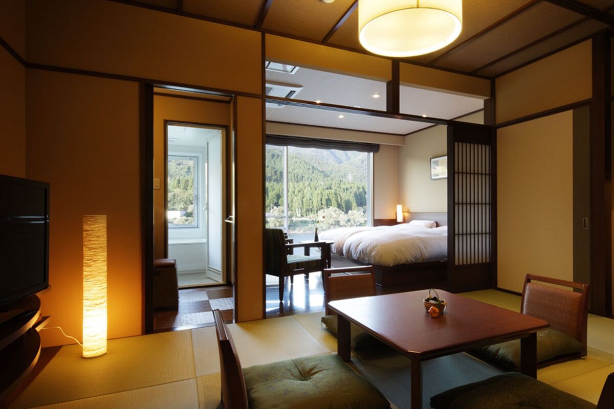 A hotel room with a bed, a dining table, chairs, a TV, and a view of a forest through the window.