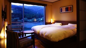 A hotel room with a large bed, a chair, a small table, and a view of a mountainous landscape through a large window.