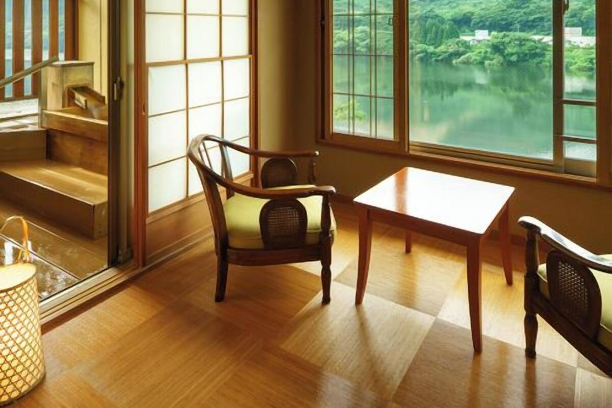 Una habitación con vistas a un lago y montañas, suelo de madera, una mesa y una silla.