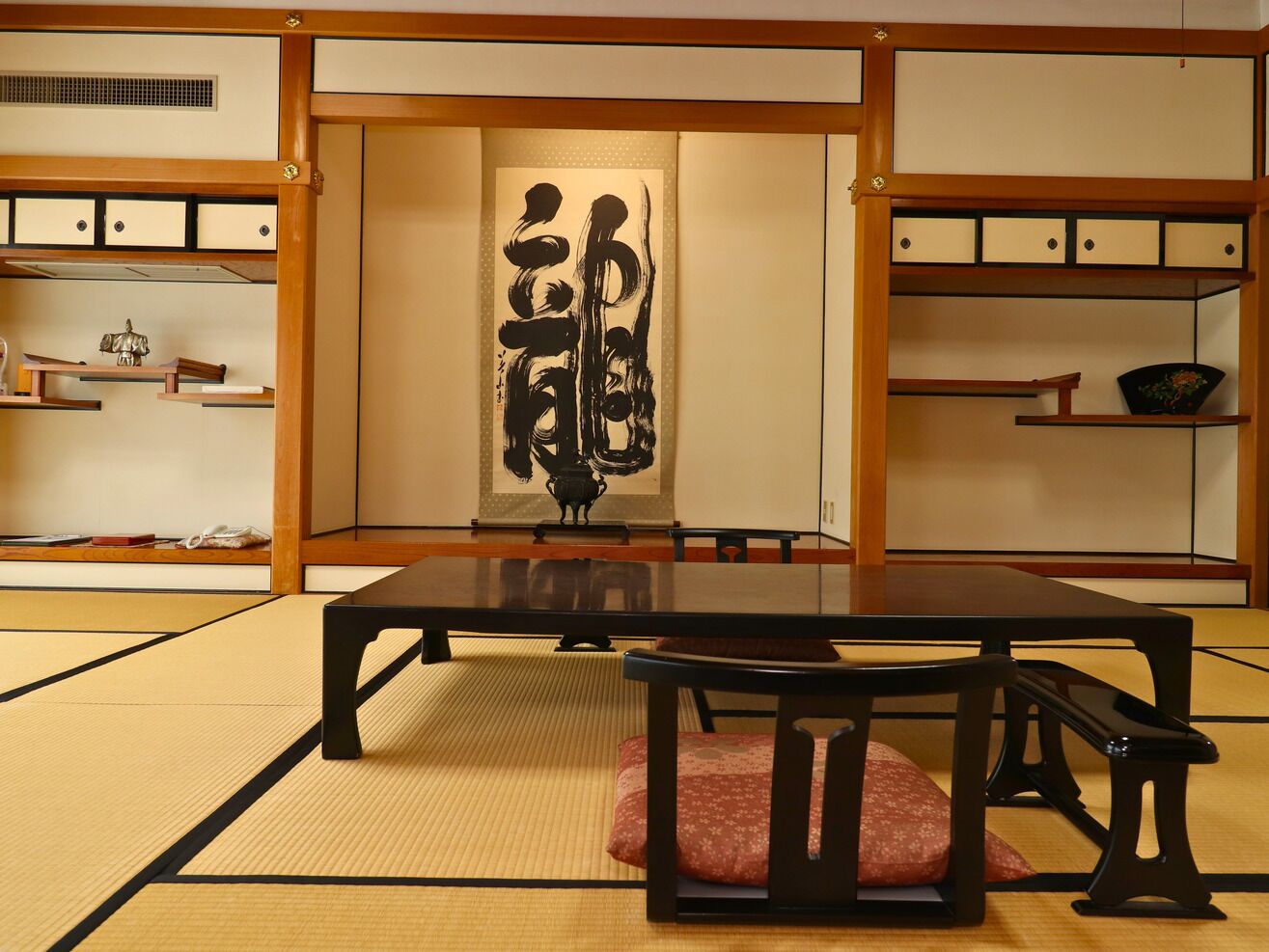 Una habitación con esteras de tatami, una mesa baja, sillas, armarios de madera y una obra de arte de caligrafía en la pared.