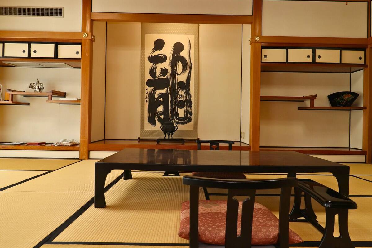 Una habitación con esteras de tatami, una mesa baja, sillas, armarios de madera y una obra de arte de caligrafía en la pared.