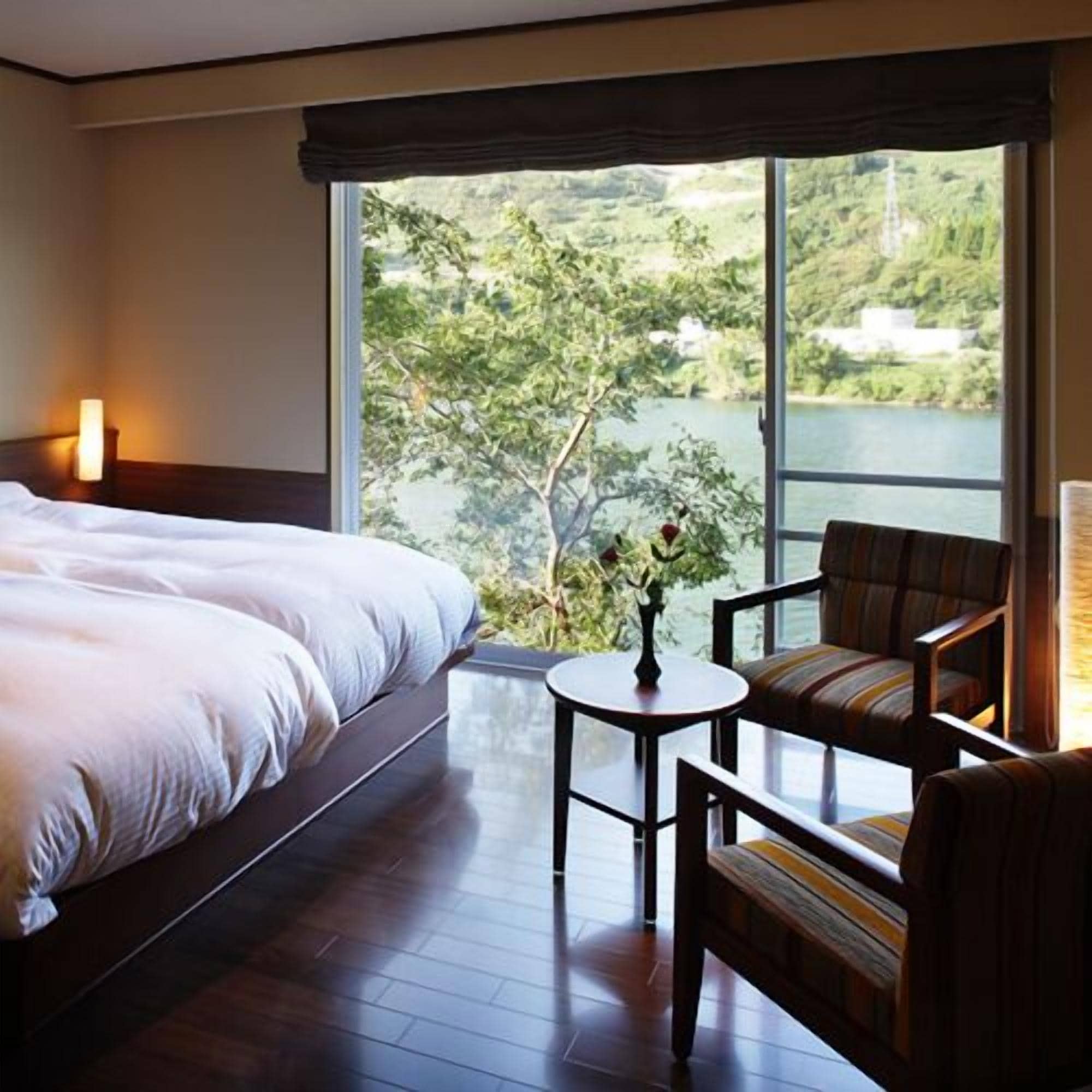 A hotel room with a large bed, a small table, a chair, and a view of a lake and greenery.