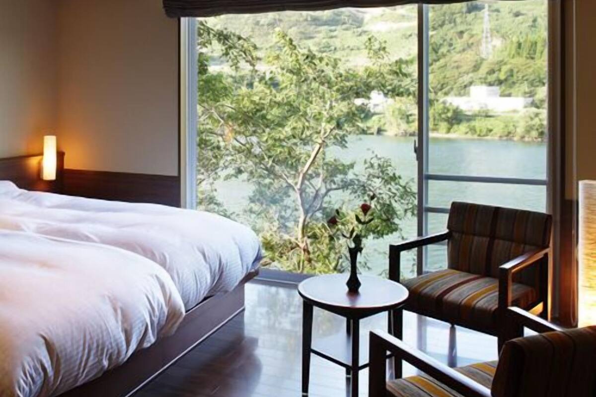 Habitación de hotel con una cama grande, una mesita, una silla y vistas a un lago y vegetación.