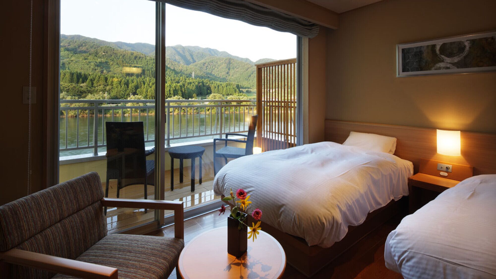 A hotel room with a bed, a chair, a small table with a vase of flowers, and a balcony with a view of a lake and mountains.