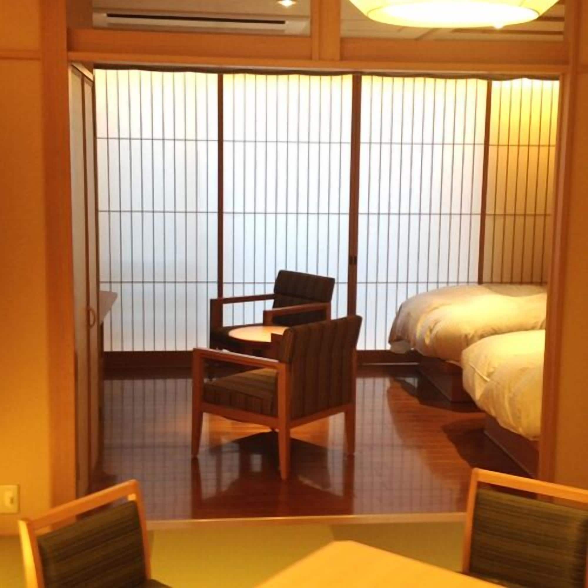A traditional Japanese room with tatami flooring, a low table with chairs, a bed with white bedding, and shoji screens.
