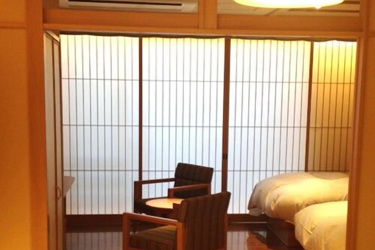 Una habitación japonesa tradicional con suelo de tatami, una mesa baja con sillas, una cama con ropa de cama blanca y puertas correderas shoji.