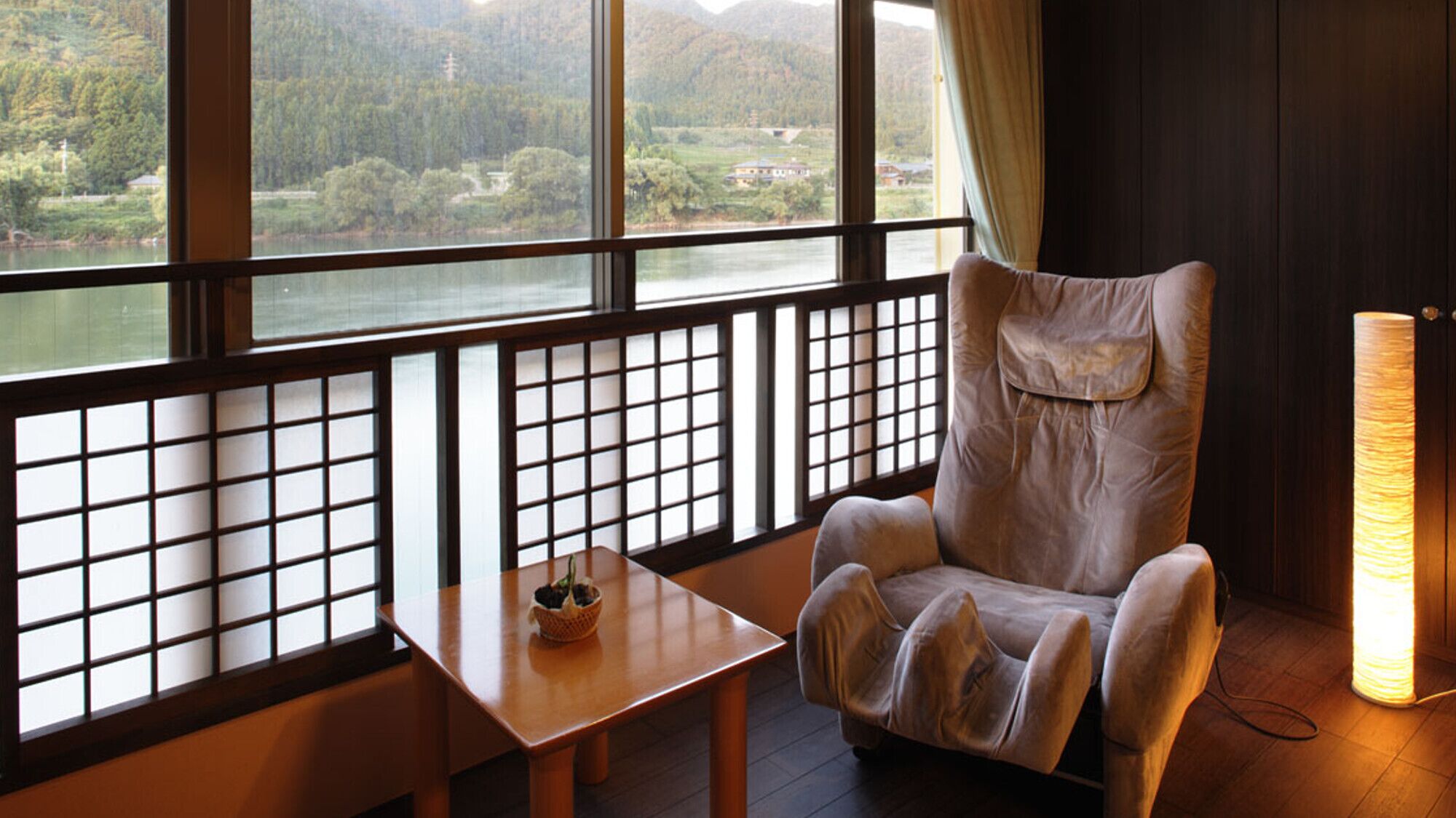 Una habitación con vistas a un lago y montañas, un sillón acolchado, una mesita con una cesta y una ventana al estilo tradicional japonés.