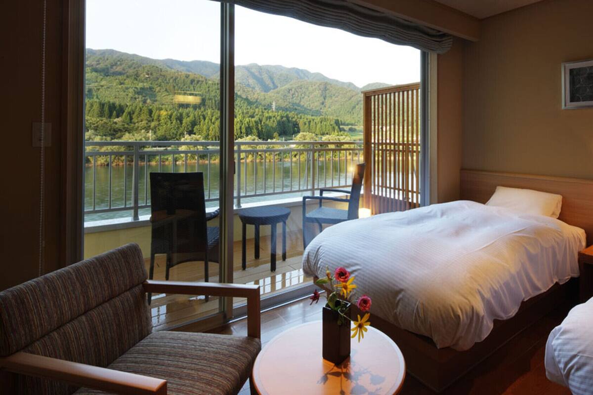 Una habitación de hotel con una cama, una silla, una mesita y vistas a un lago y montañas.