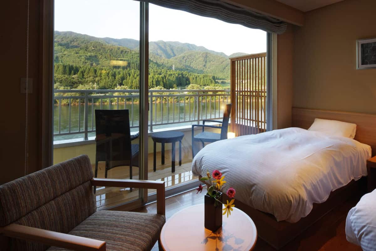 Una habitación de hotel con una cama, una silla, una mesita y vistas a un lago y montañas.