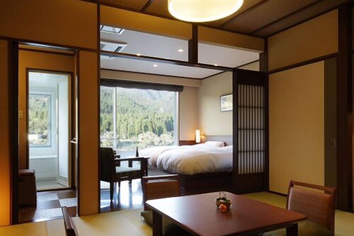 Una habitación tradicional japonesa con suelo de tatami, una mesa baja, cojines y una cama con vistas al exterior.