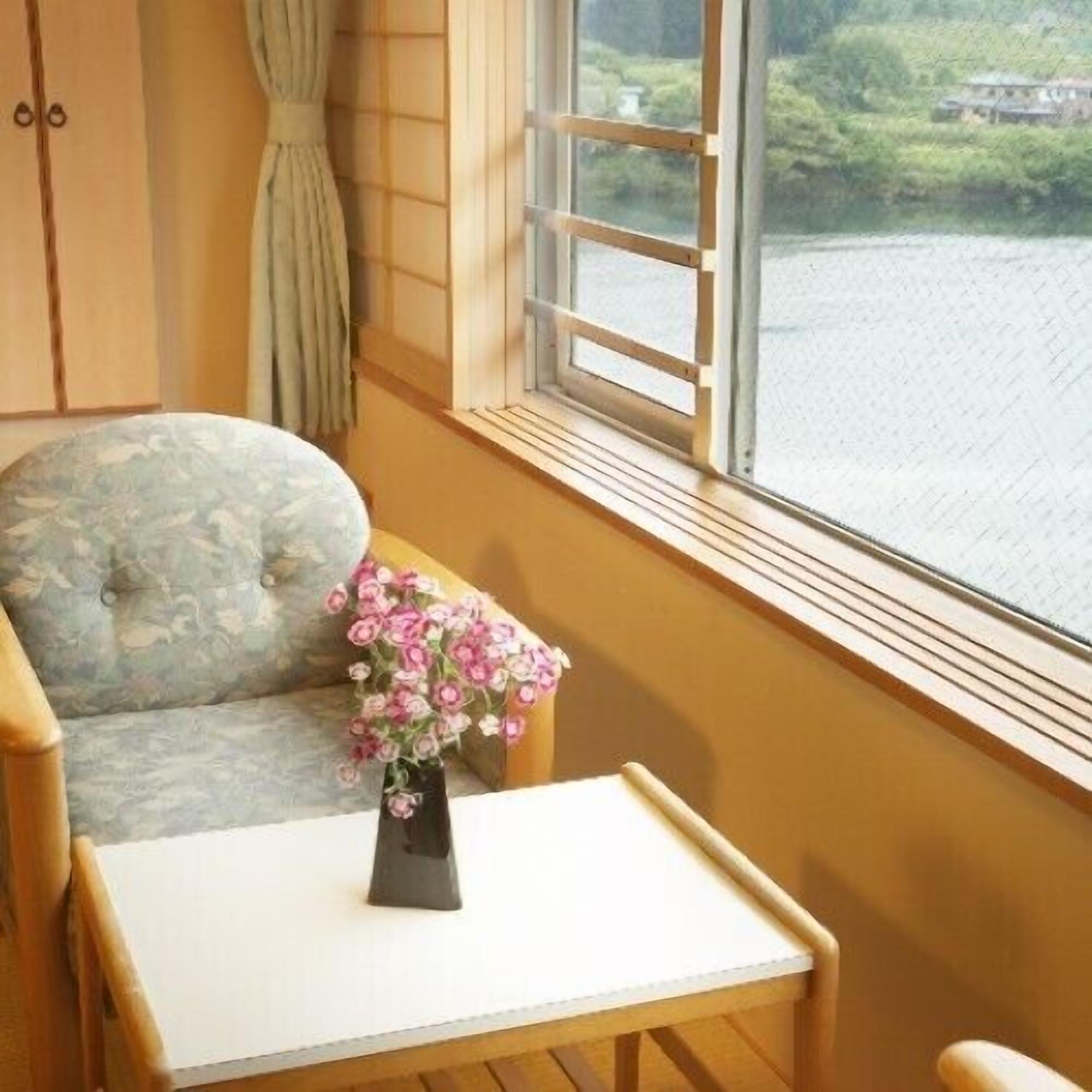 Una habitación con ventana, una silla, una mesita y un florero con flores.