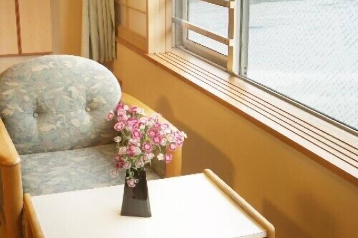 Una habitación con ventana, una silla, una mesita y un florero con flores.