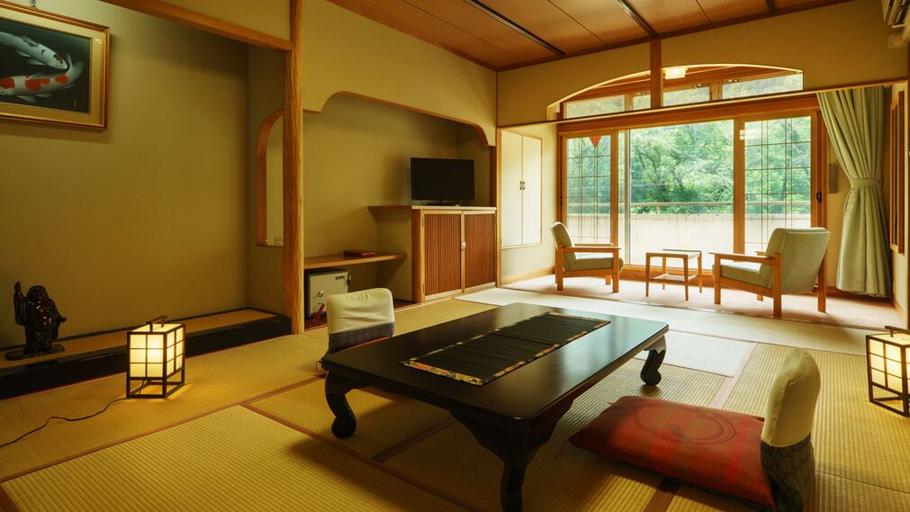 Una habitación tradicional japonesa con suelo de tatami, una mesa baja, sillas, televisión y vistas a través de amplias puertas correderas.