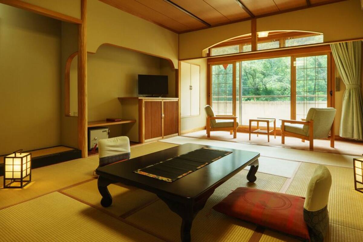 Una habitación tradicional japonesa con suelo de tatami, una mesa baja, sillas, televisión y vistas a través de amplias puertas correderas.
