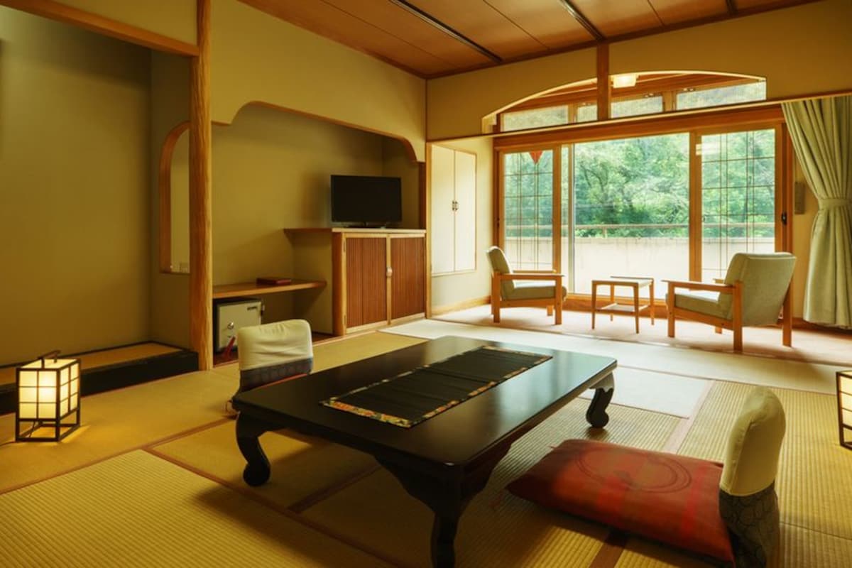 Una habitación tradicional japonesa con suelo de tatami, una mesa baja, sillas, televisión y vistas a través de amplias puertas correderas.