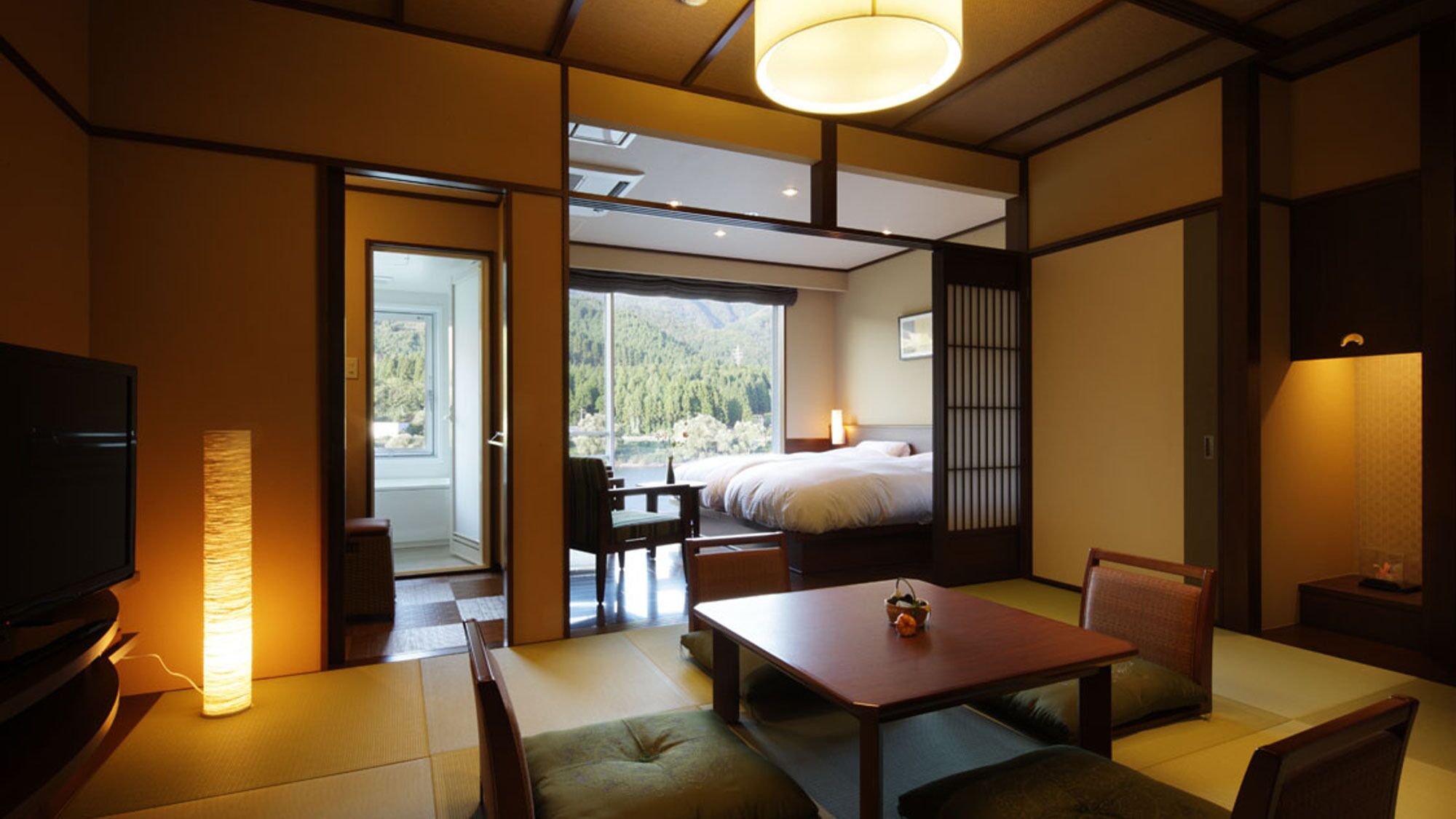 A hotel room with a bed, a dining table, chairs, a TV, and a view of a forest through the window.