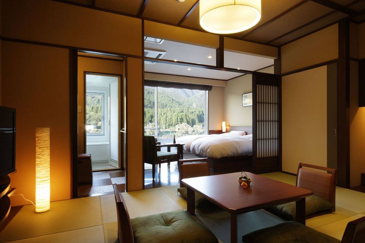 Habitación de hotel con cama, una mesa de comedor, sillas, televisor y vista al bosque a través de la ventana.