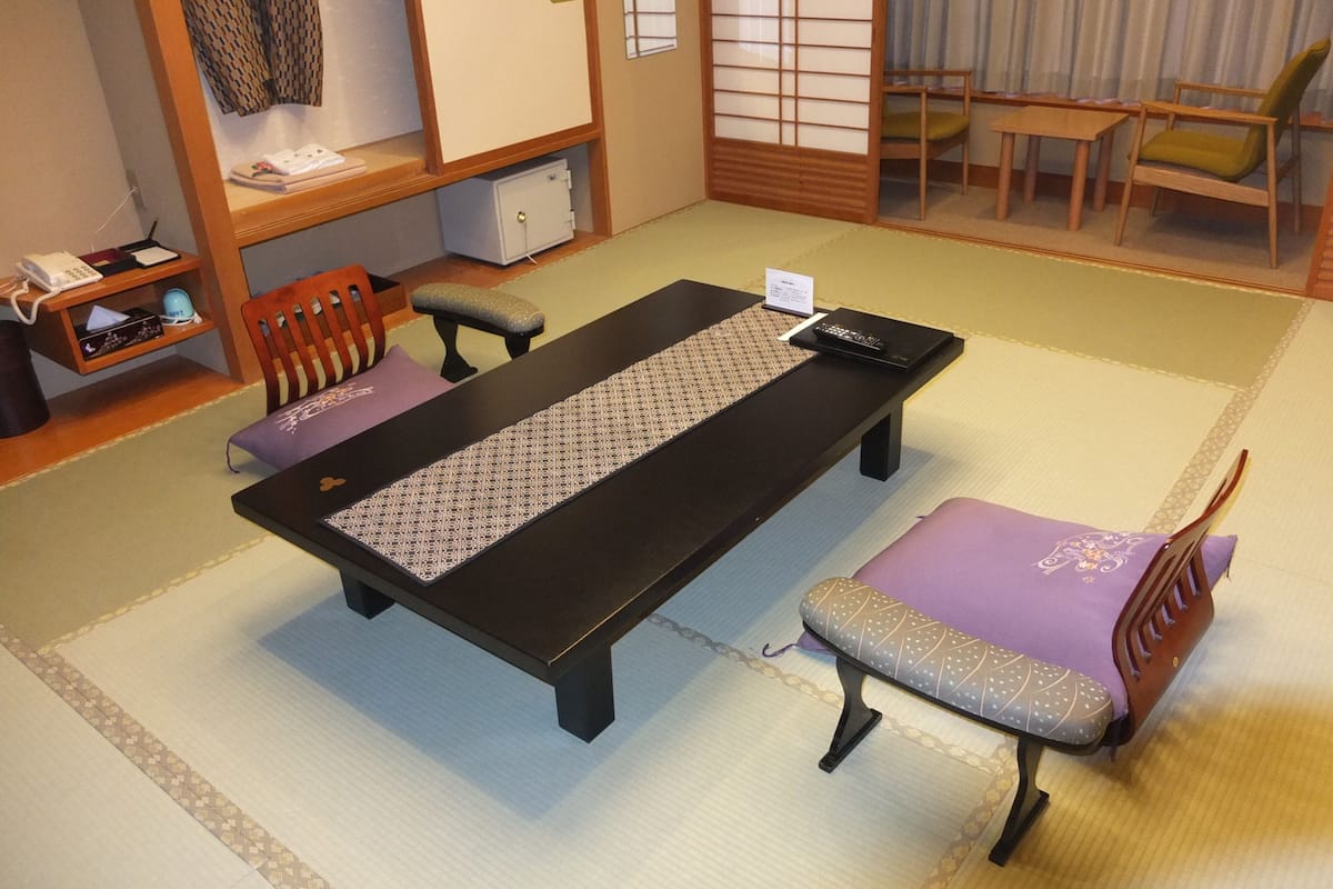 Una habitación con esteras de tatami, una mesa baja, dos sillas, un armario pequeño y una ventana con puertas japonesas shoji.