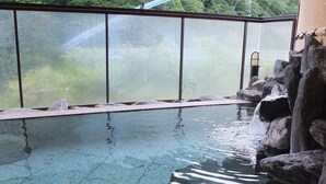 An outdoor swimming pool with clear water, surrounded by rocks and a view of a forested area and a bridge. - Kashiwaya (Nikko)