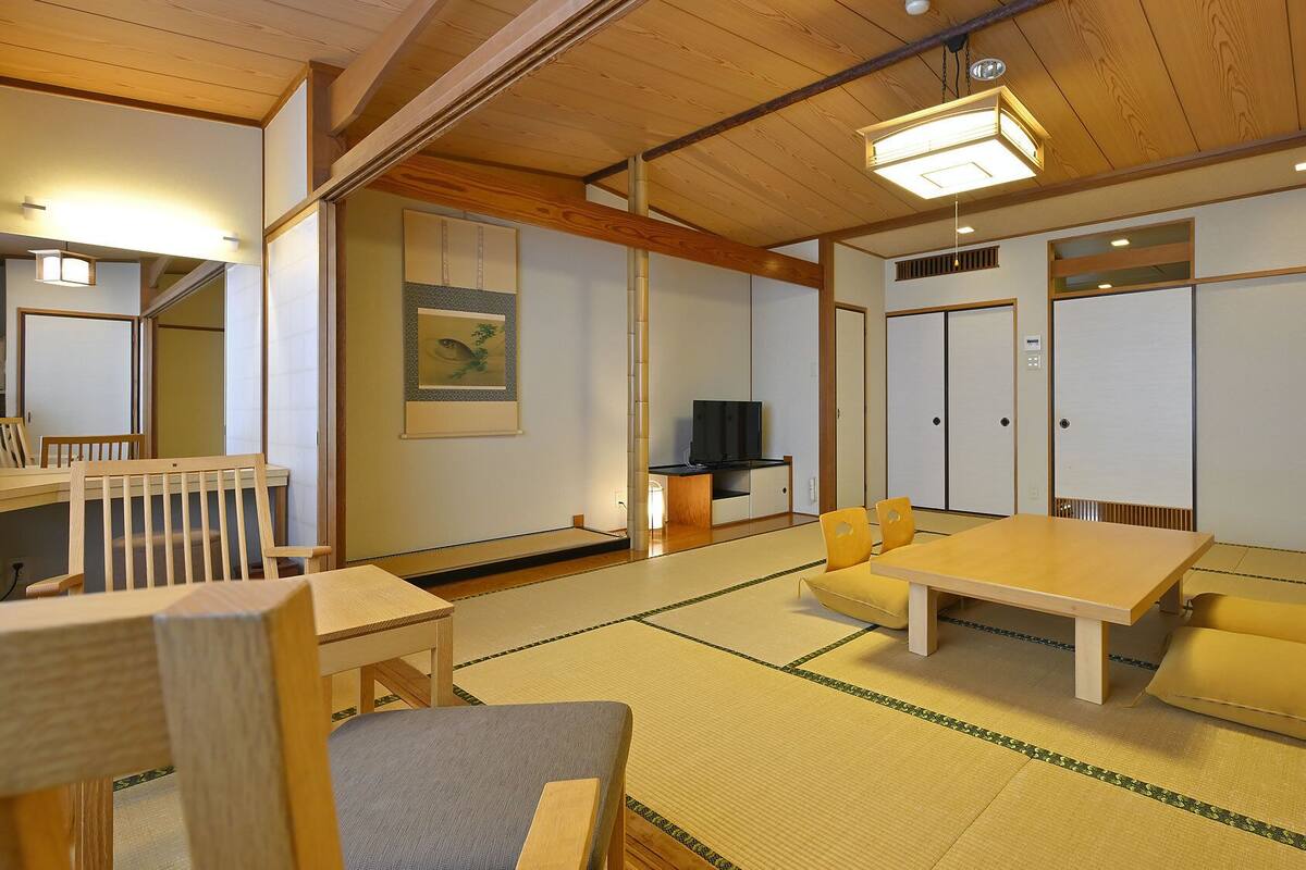 Une pièce de style japonais traditionnel, avec un sol recouvert de tatamis, une table basse, des chaises, un téléviseur et des portes coulissantes.