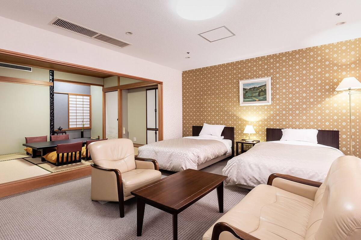 Une chambre d’hôtel avec un lit, deux chaises, une petite table, un bureau, un miroir, un tableau au mur et une fenêtre.