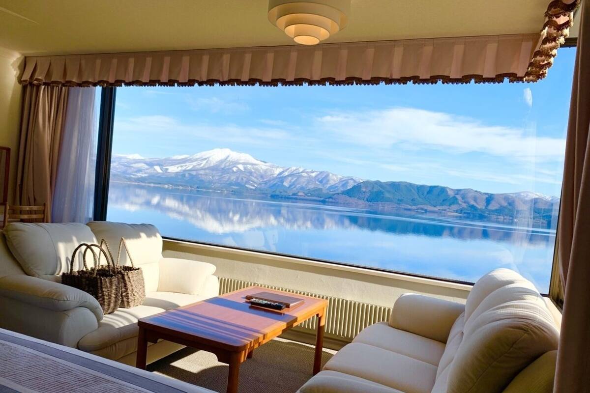 Ein Zimmer mit großem Fenster, einer Couch, einem Tisch und Blick auf einen See und Berge.