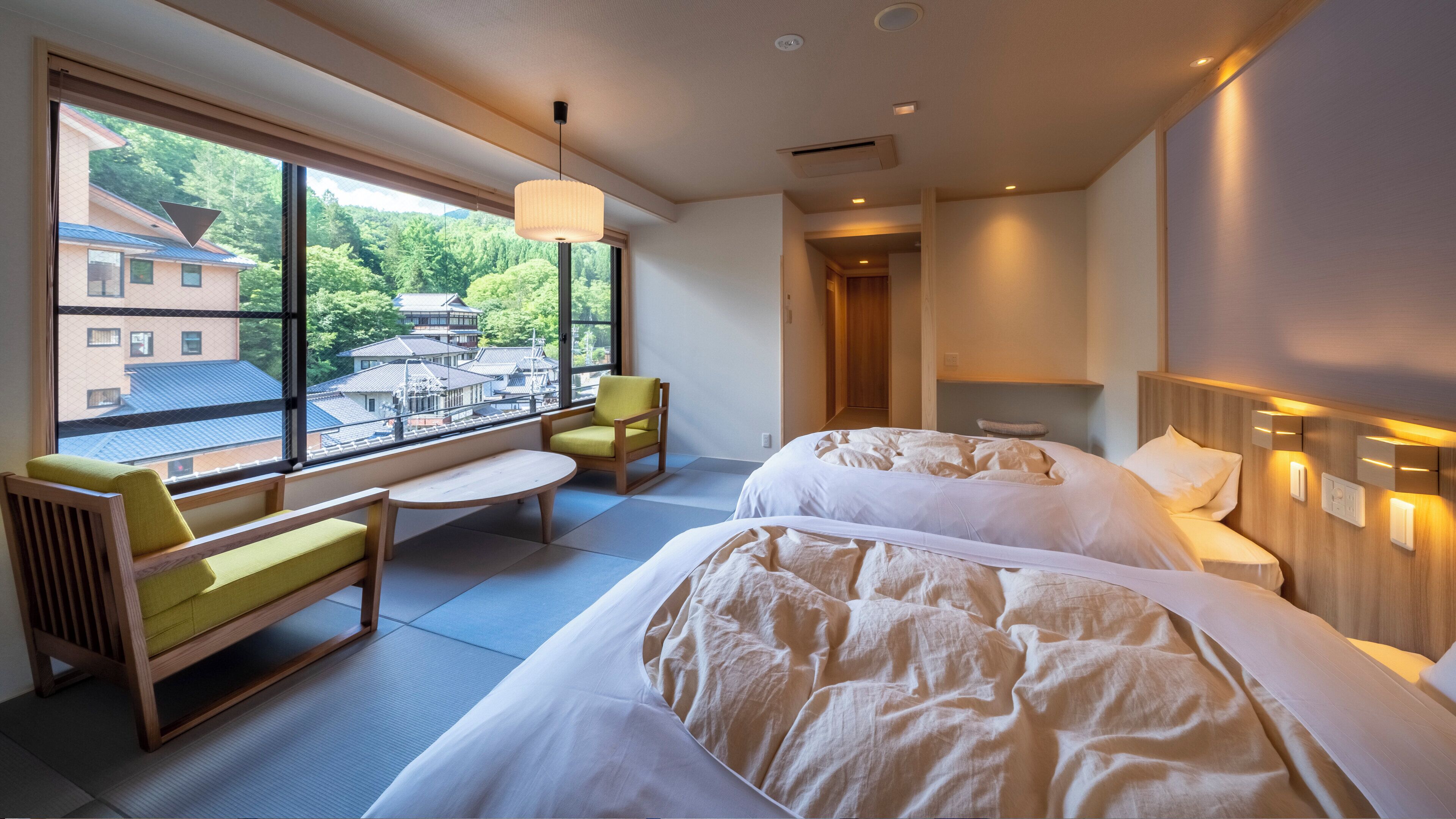 A hotel room with two beds, a sofa, a chair, and a large window offering a view of buildings and trees.