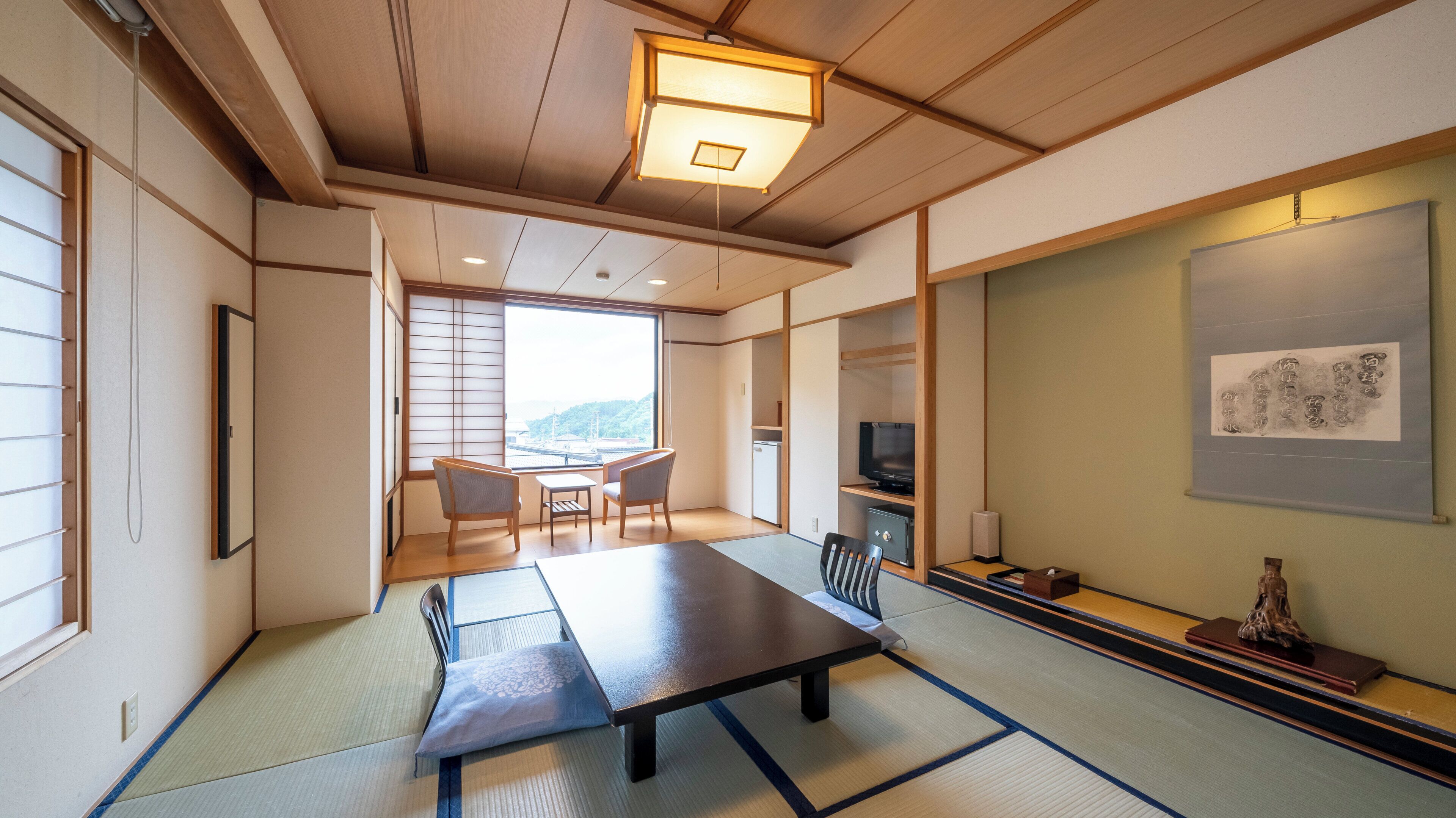 A traditional Japanese room with tatami flooring, a low table, chairs, a television, and a view of a mountain.