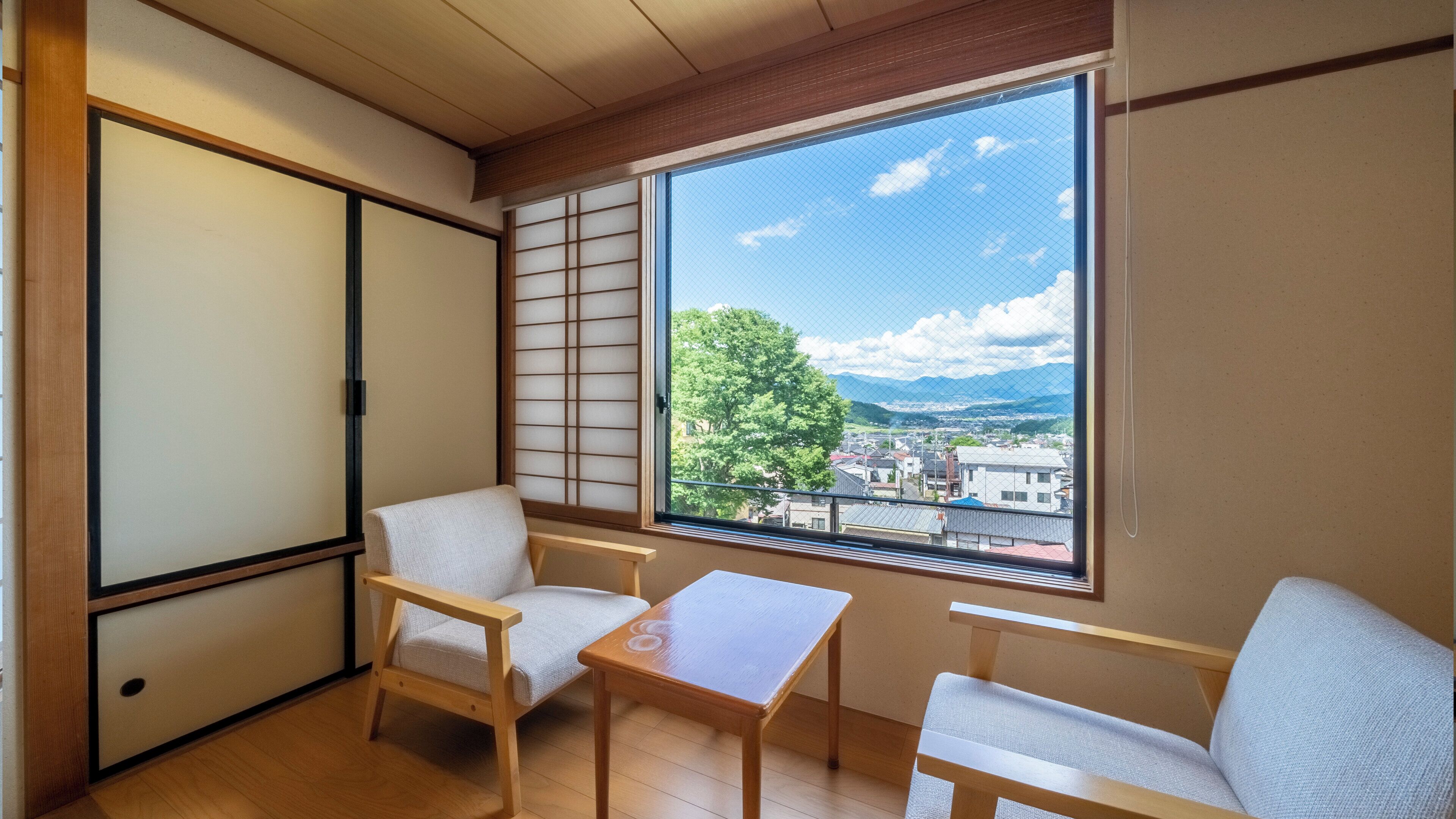 A room with tatami flooring, a sliding shoji screen, a wooden table, two chairs, and a large window with a view of a tree and buildings.